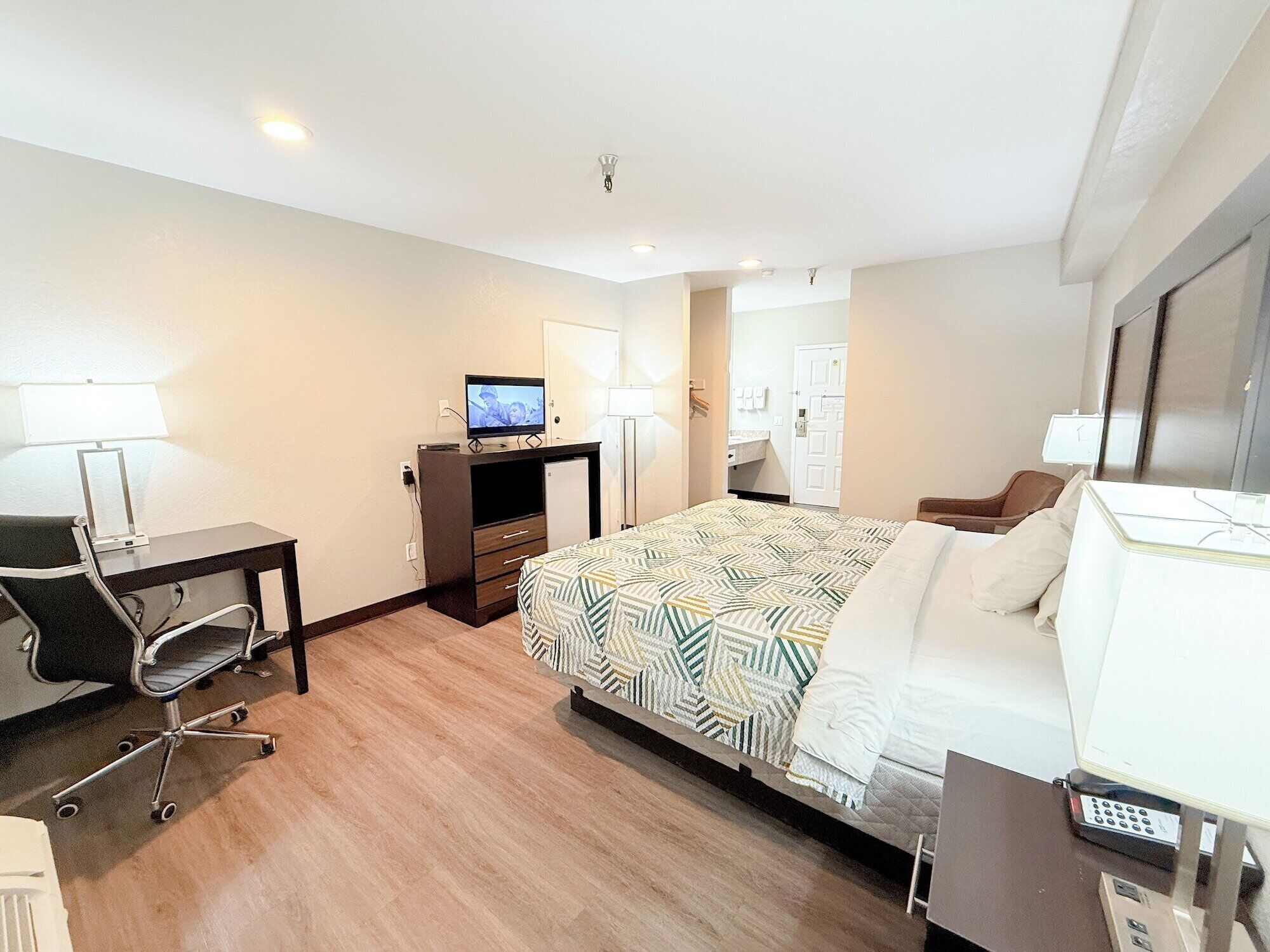 A hotel room with a bed, a desk with a computer, a chair, a TV, and a bathroom visible through an open door.