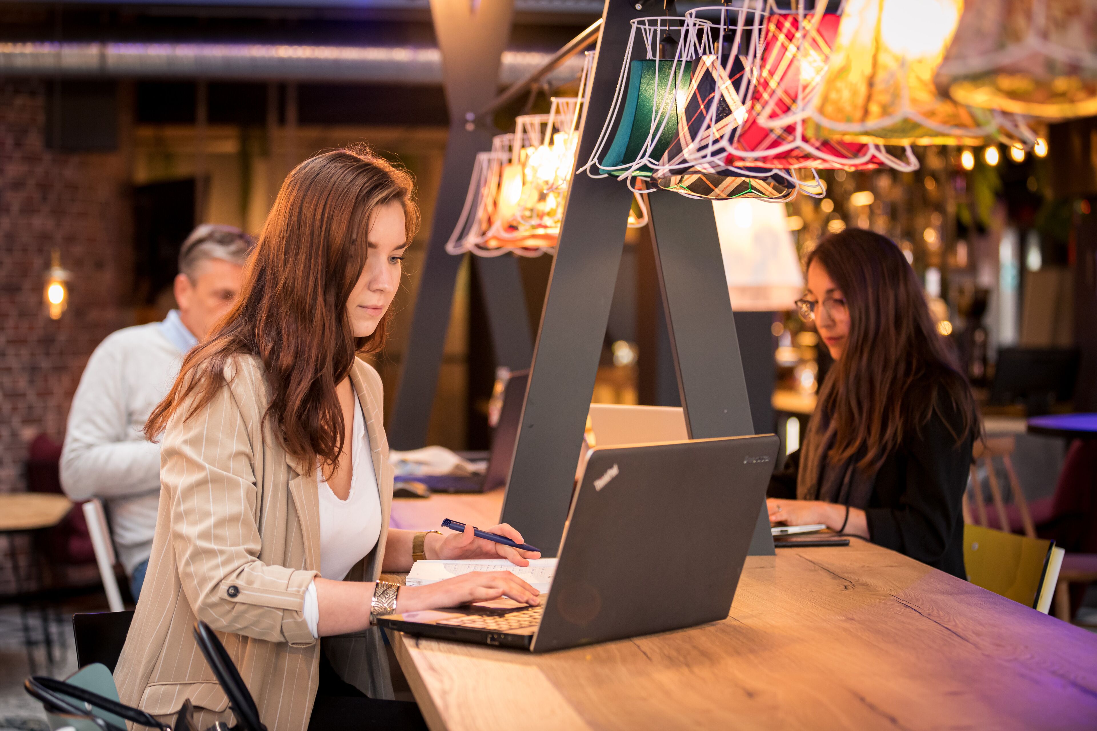 A person working on a laptop at a wooden table in a cafe with a brick wall and hanging lights in the background.