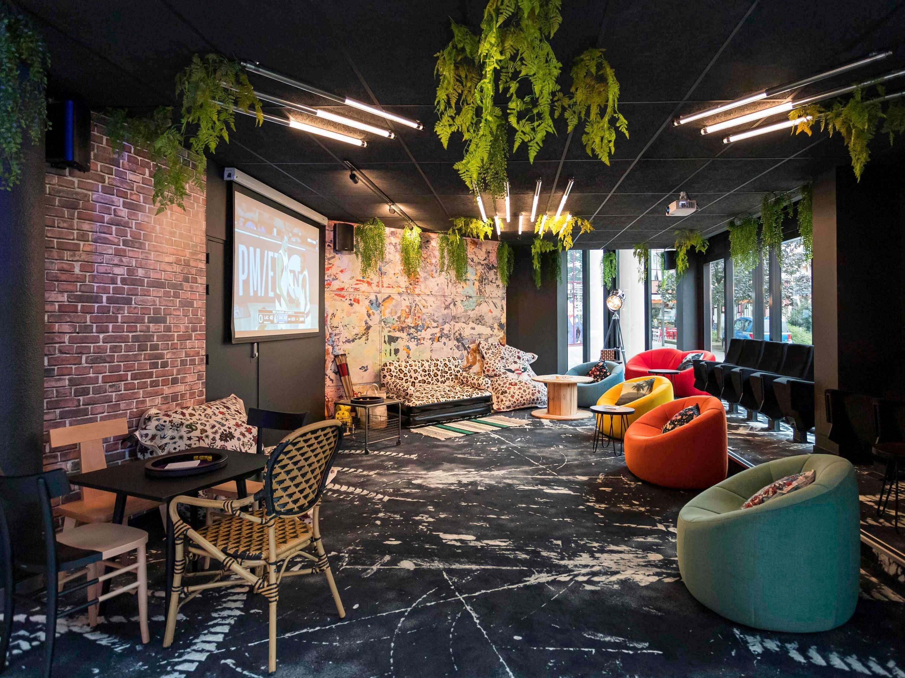 A modern lounge area with a brick wall, a projector screen, a sofa with a leopard print cover, and various seating options including a red chair and a teal bean bag.