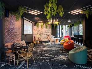 A modern lounge area with a brick wall, a projector screen, a sofa with a leopard print cover, and various seating options including a red chair and a teal bean bag. - ibis Styles Strasbourg Nord Palais des Congrès (Schiltigheim)