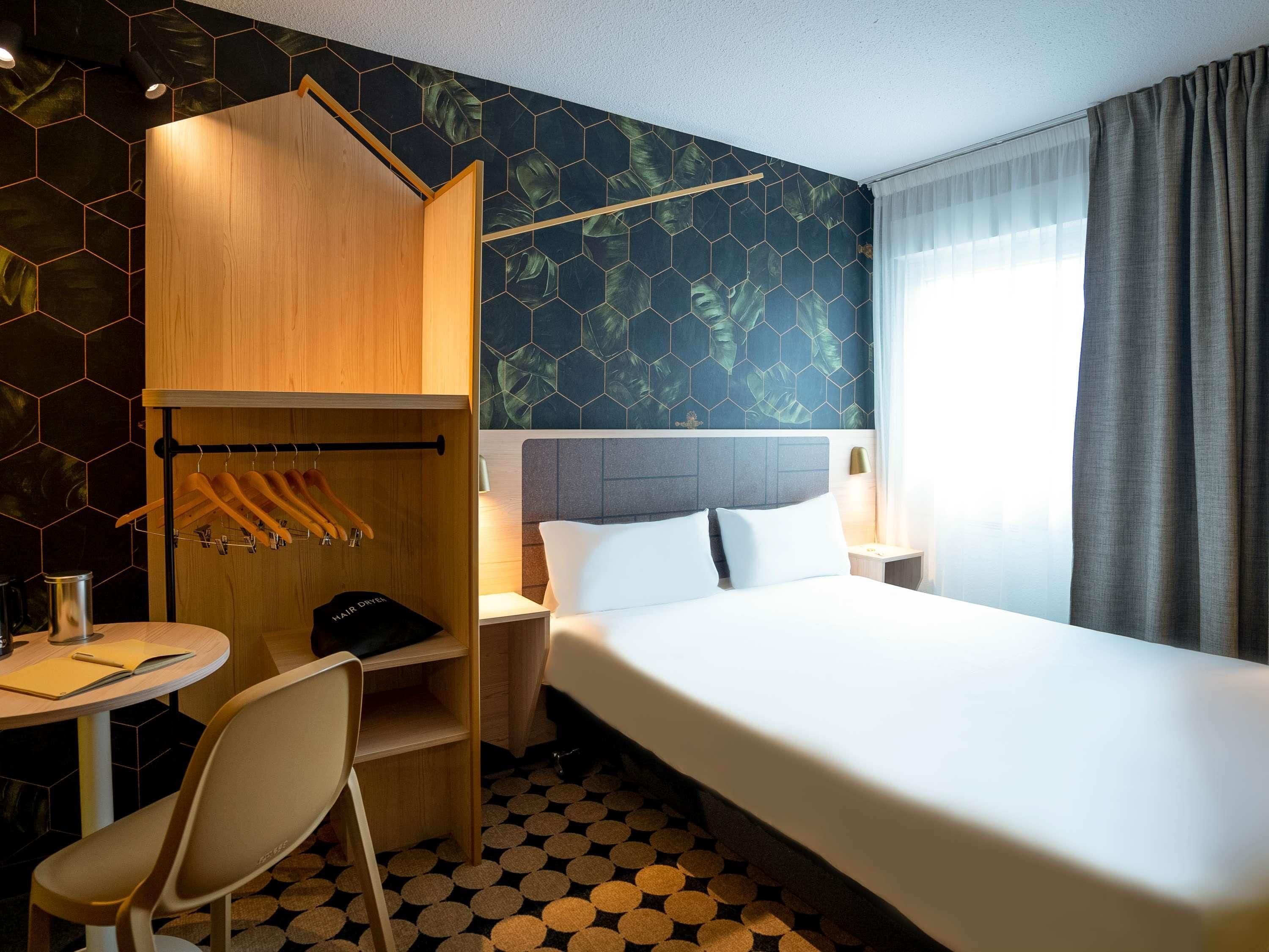 A hotel room with a bed, a wooden wardrobe, a desk with a chair, and a round table.