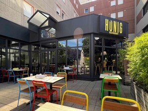 Ulkoterassi, jossa on pöytiä ja tuoleja, lasiseinäinen tila ja rakennus, jonka yläpuolella on kyltti, jossa lukee: ”HUNIG RESTAURANT”.