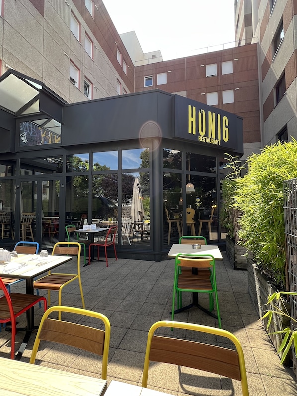 Un área de asientos al aire libre de un restaurante, con mesas y sillas, rodeada de edificios.