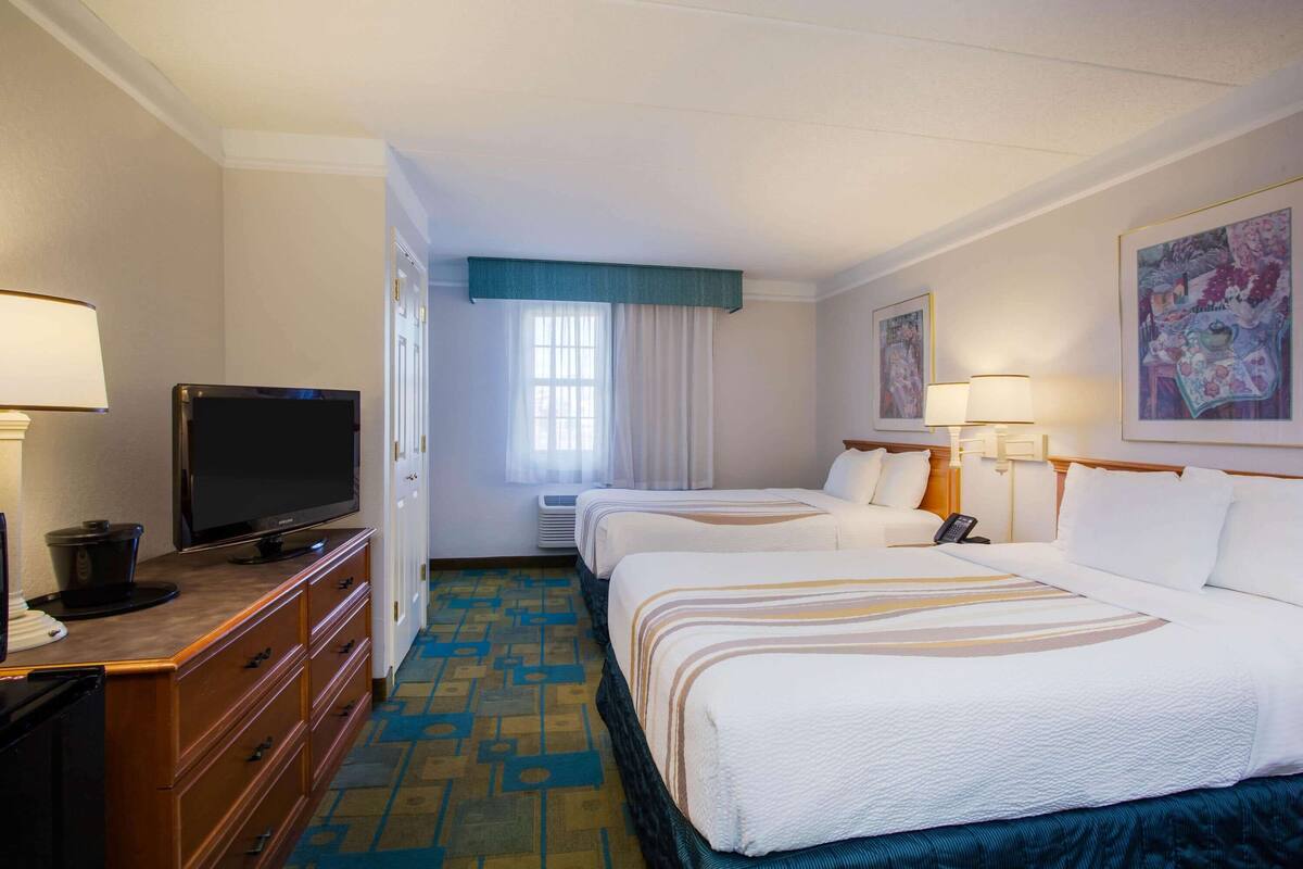 A hotel room with a television on a wooden dresser, two beds with white bedding, a blue carpet, and a painting on the wall.