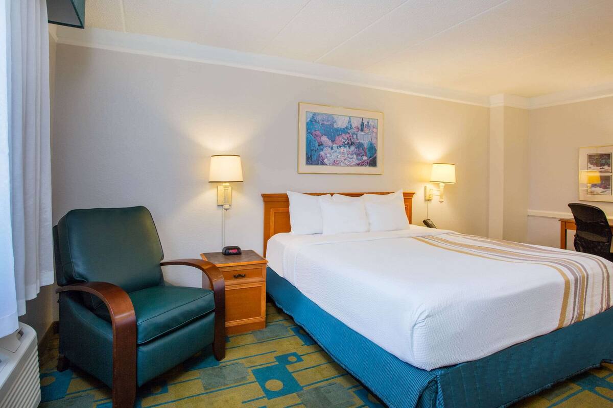 A hotel room with a bed, a chair, a nightstand, a lamp, and a painting on the wall.
