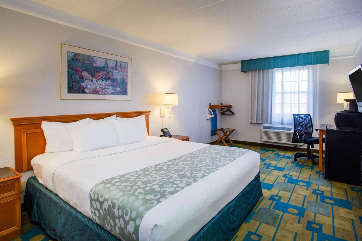 A hotel room with a large bed, a desk with a chair, a window with curtains, and a painting on the wall.