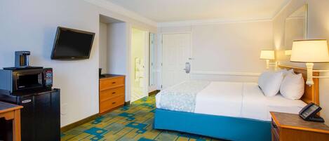 A hotel room with a bed, a desk with a microwave, a TV, and a bedside table with a lamp.