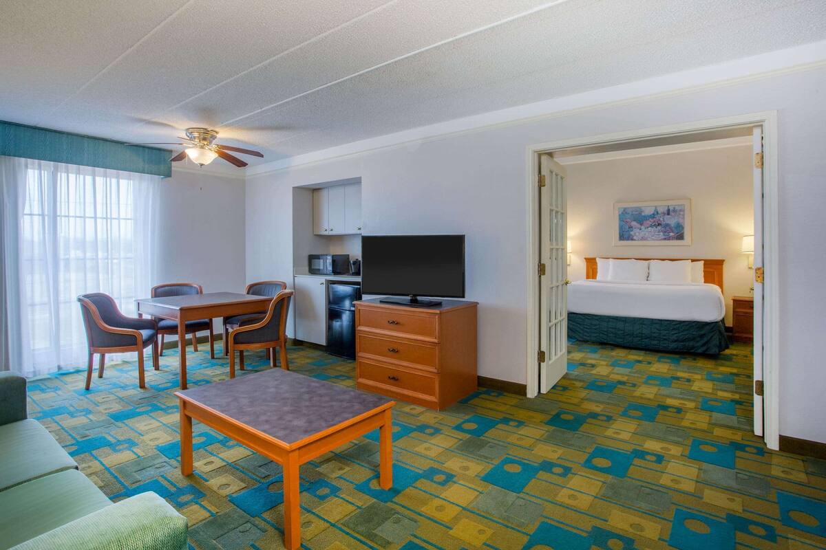 A hotel room with a bed, a desk with a television, a dining area with chairs, and a patterned carpet.