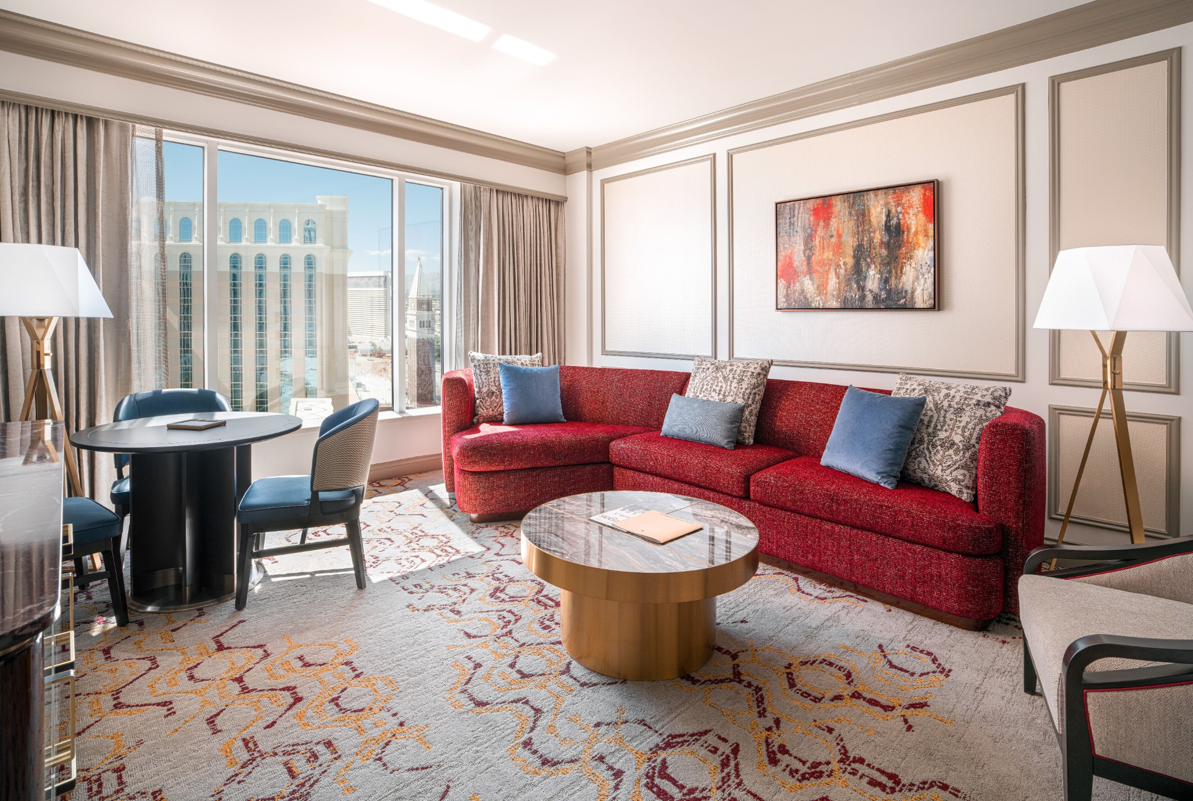 A hotel room with a red sofa, a round coffee table, a dining table with chairs, and a large window with curtains.