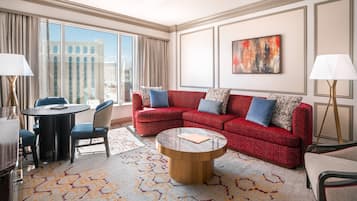 A hotel room with a red sofa, a round coffee table, a dining table with chairs, and a large window with curtains.