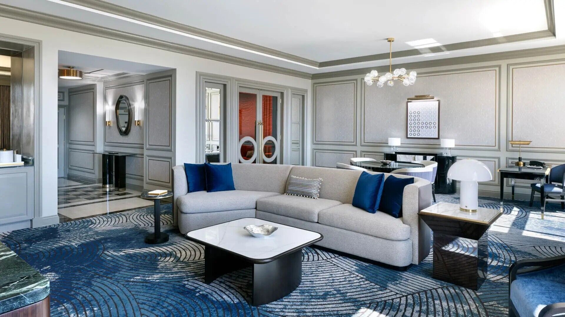 Una habitación de hotel moderna con un sofá gris, una mesa de centro blanca y una alfombra azul estampada.
