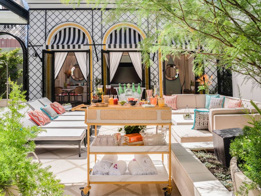 Uma área externa iluminada pelo sol, com cadeiras de lounge, uma mesa de rattan com comodidades, e um edifício ao fundo com arcos e uma parede de azulejos.