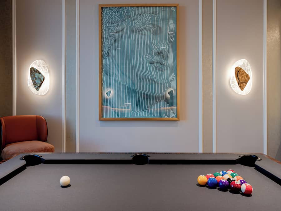 Una mesa de billar con una bola blanca y bolas multicolores, un cuadro abstracto enmarco, y dos apliques de pared iluminados.