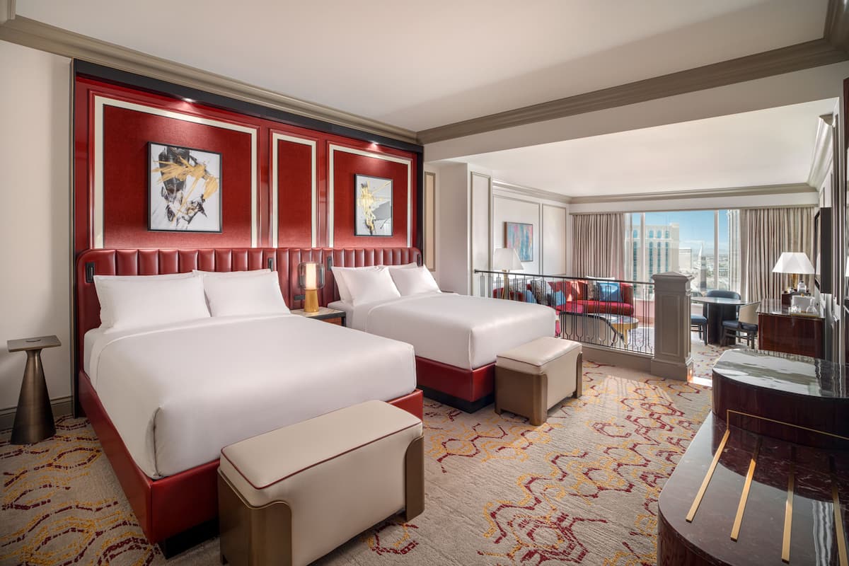 A hotel room with two beds, a sitting area, a balcony with a view, and a desk with a lamp.