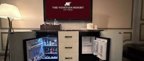 A minibar with a TV displaying 'The Venetian Resort Las Vegas' logo, a lamp, and a refrigerator with open doors revealing beverages.