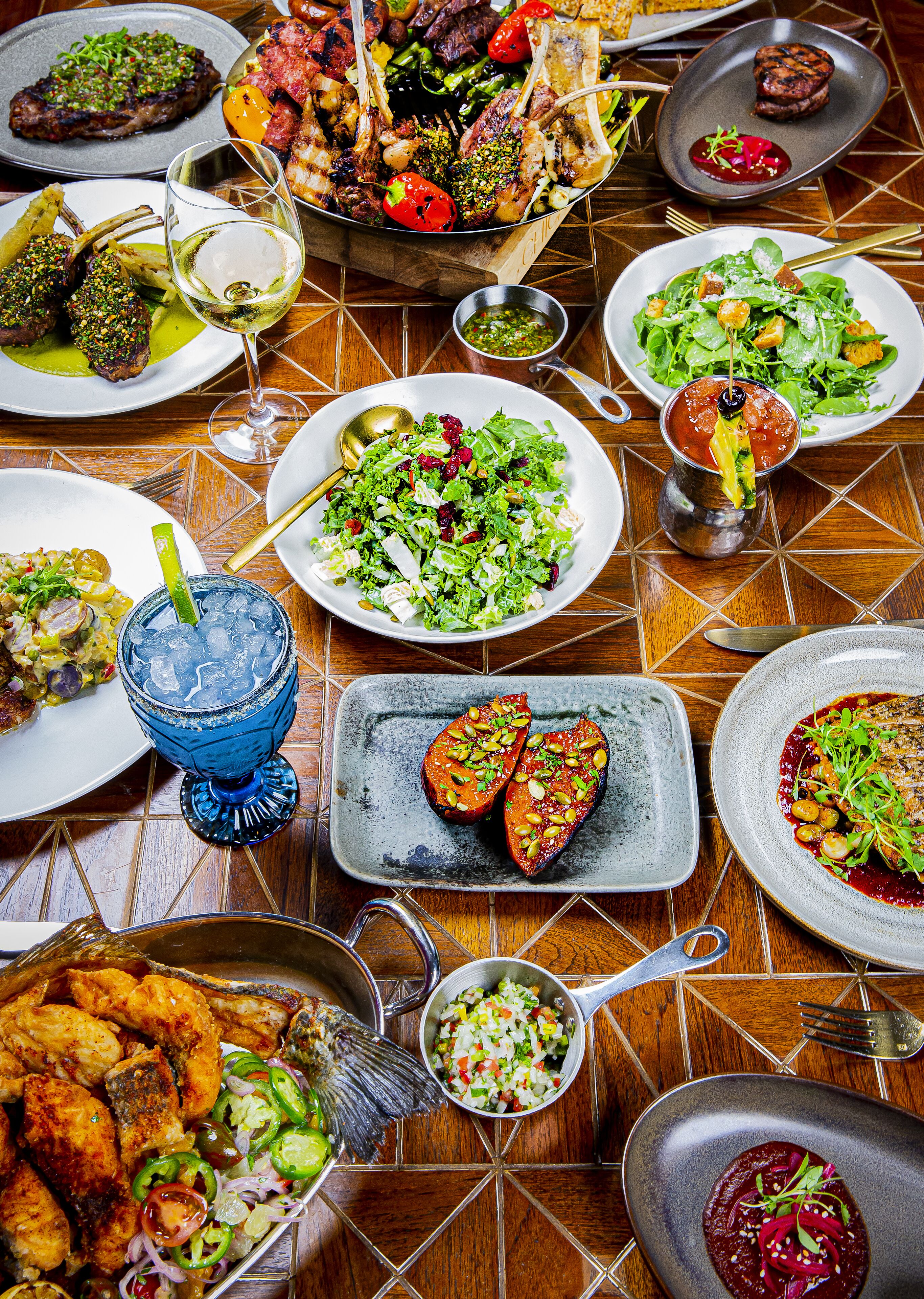 A variety of dishes on a wooden table, including grilled meats, salads, and a blue cocktail glass.
