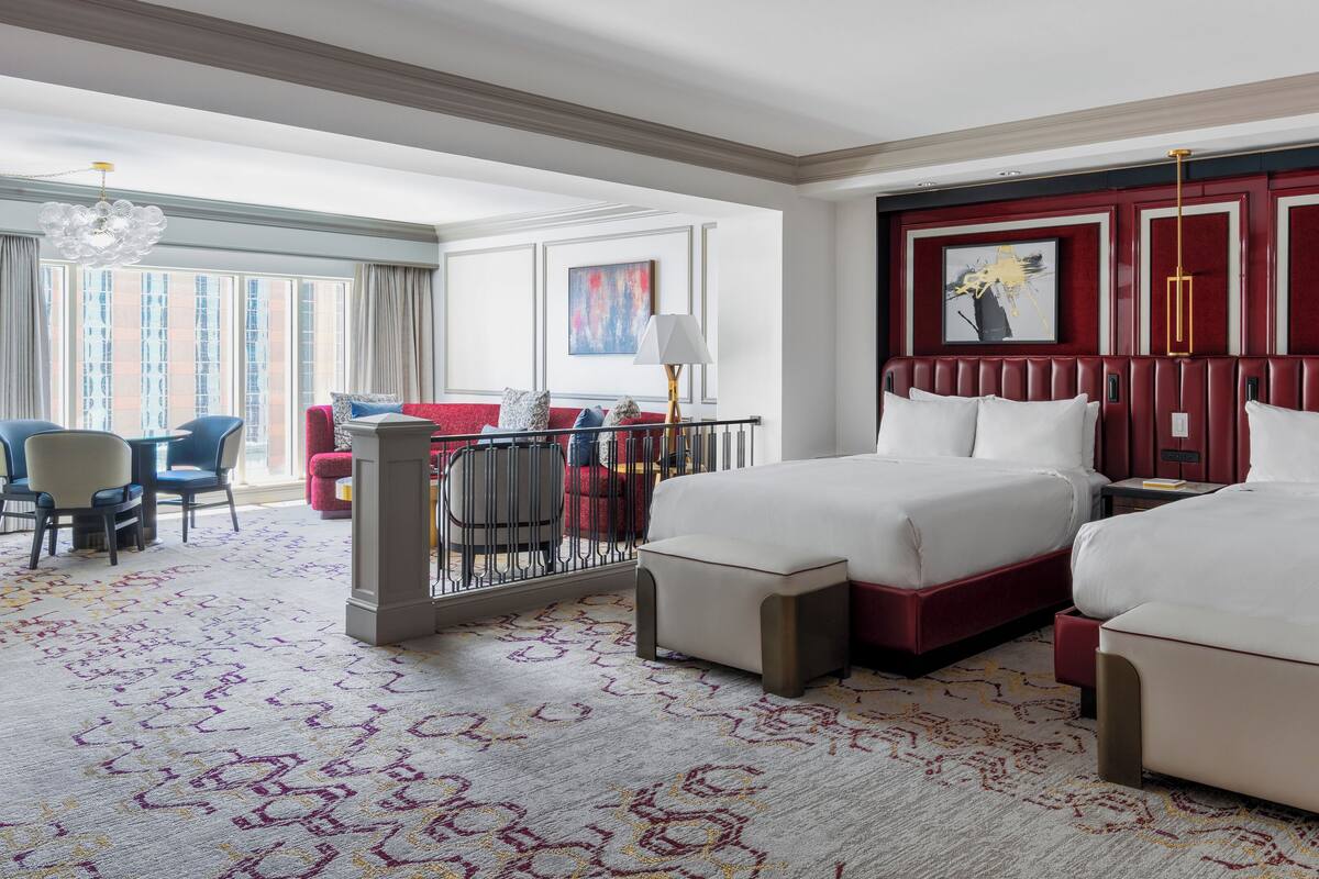 A hotel room with two beds, a sitting area with a red sofa and blue chairs, a large window with curtains, and artwork on the walls.