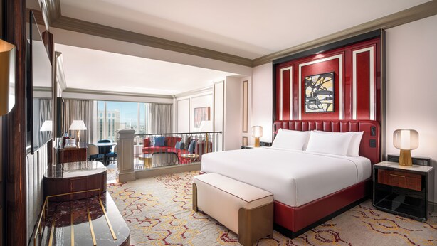 A hotel room with a large bed, a sitting area with a chair, a desk, and a large window offering a city view.