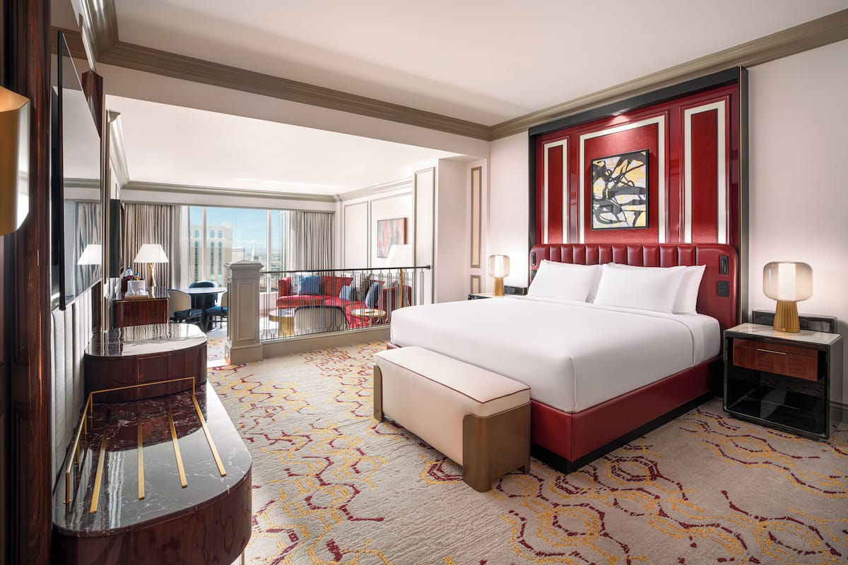 A hotel room with a large bed, a sitting area with a chair, a desk, and a large window offering a city view.