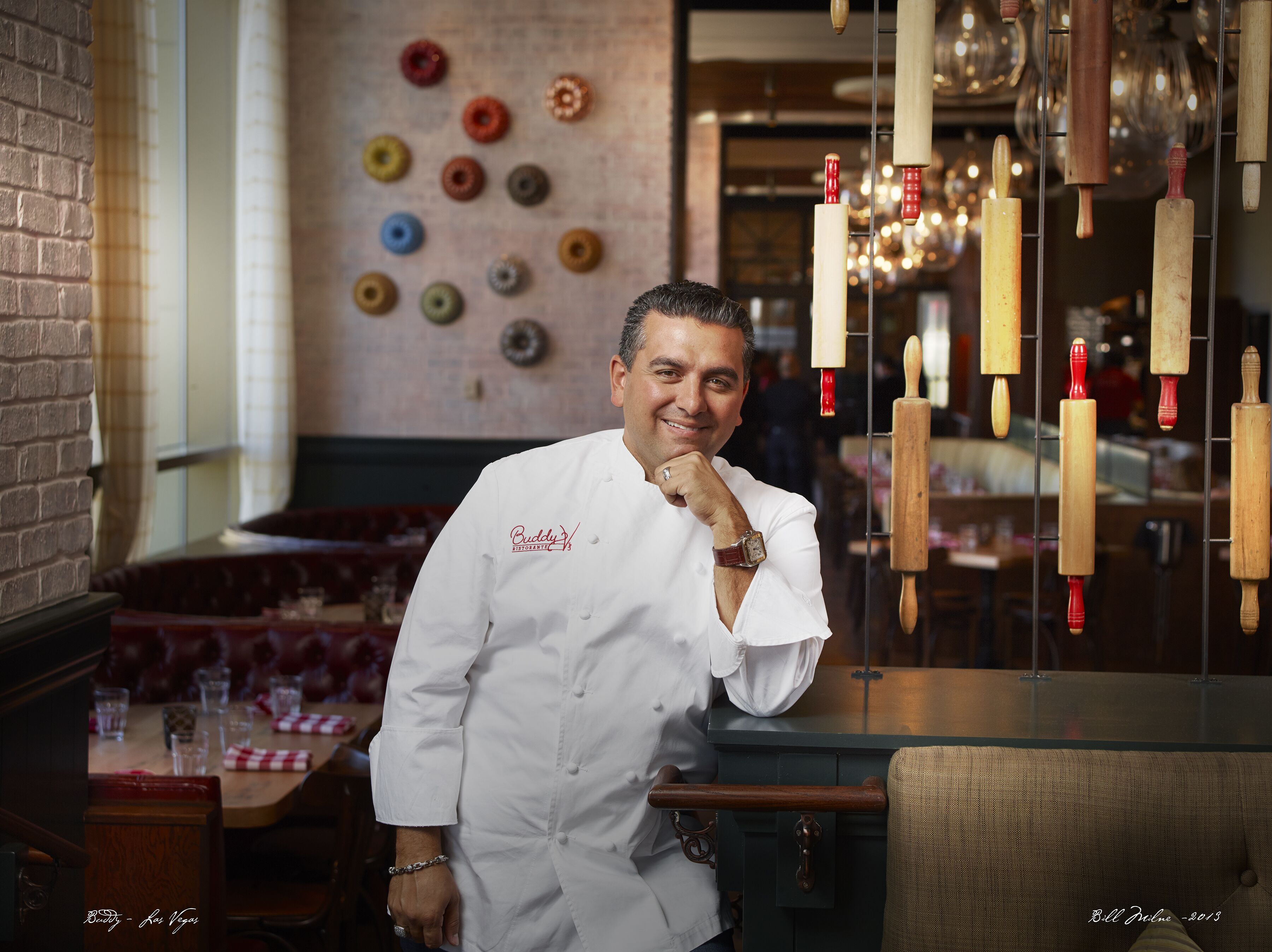A chef in a white uniform with a watch, standing in a restaurant with a brick wall and a wall of colorful buttons.