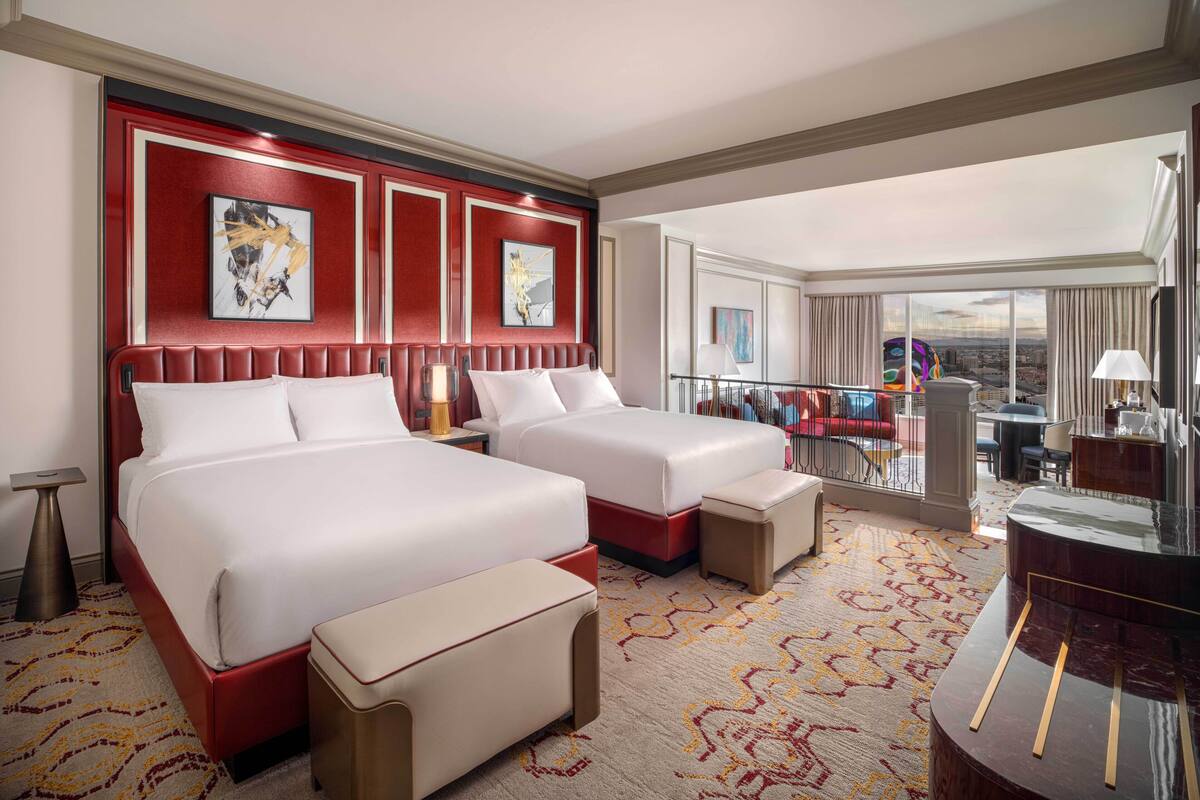 A hotel room with two beds, a desk, and a balcony with a city view.