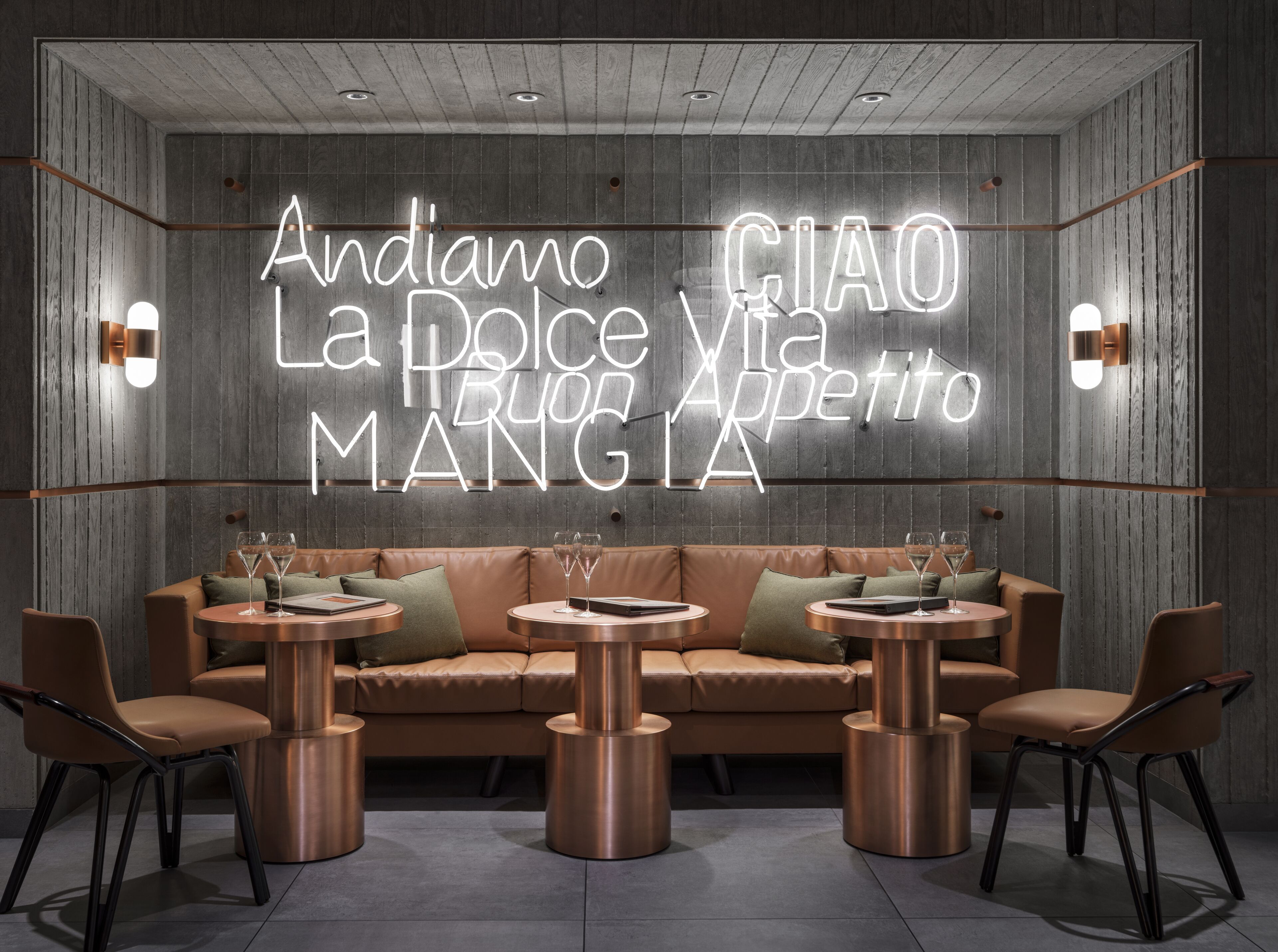 A modern interior with a neon sign reading "Andiamo La Dolce Vita Appetito Mangia" above a leather sofa and round tables.