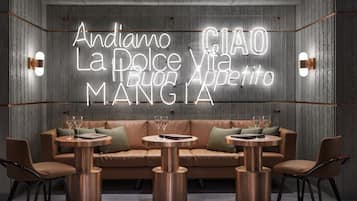 A modern interior with a neon sign reading "Andiamo La Dolce Vita Appetito Mangia" above a leather sofa and round tables.
