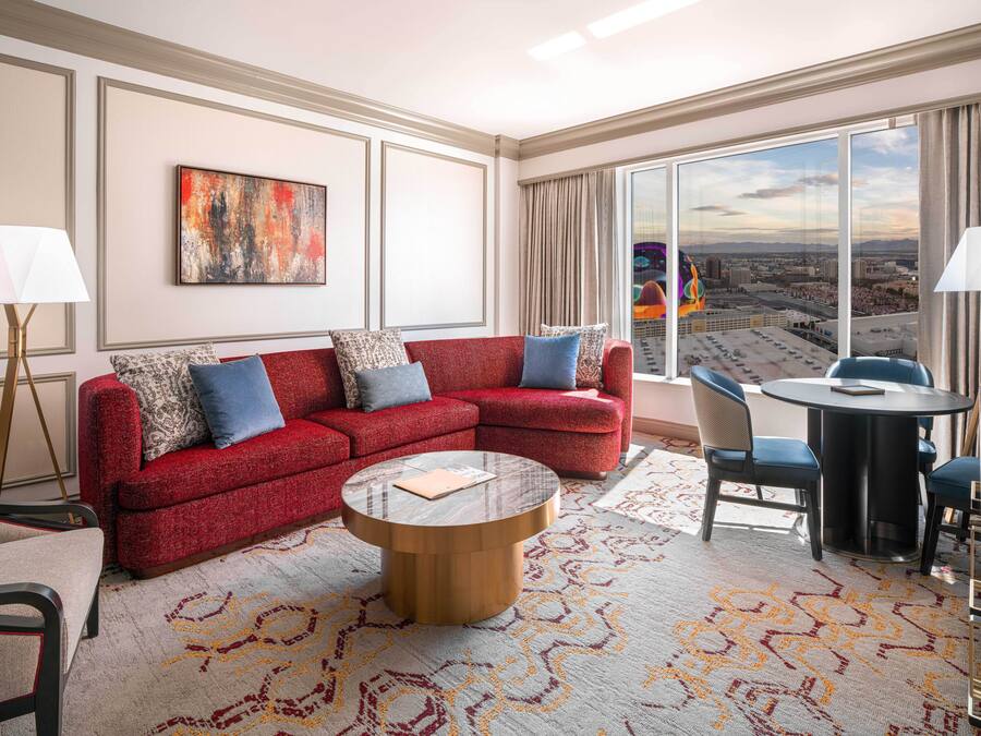 A modern hotel room with a red sectional sofa, a round coffee table, a dining area with a small table and chairs, and a large window offering a city view.