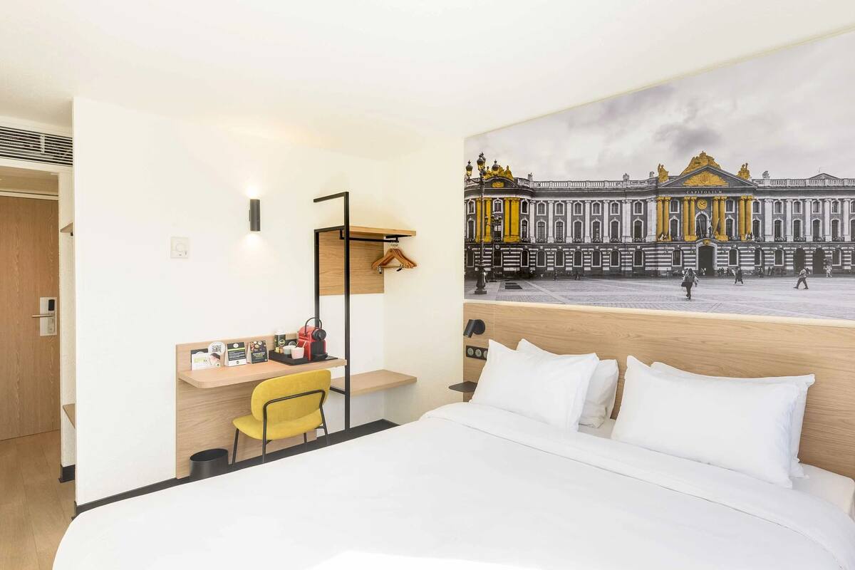 A hotel room with a bed, a desk with a chair, a wall-mounted shelf, and a large window with a view of a historic building.