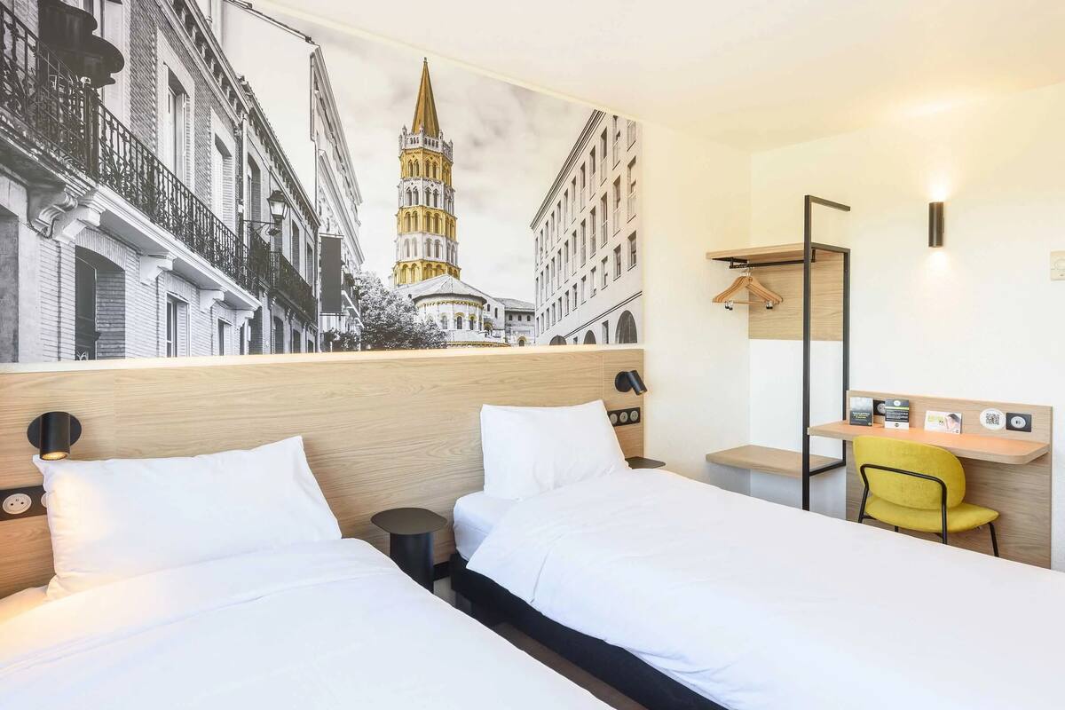 A hotel room with two beds, a desk, a chair, a coat rack, and a mural of buildings.