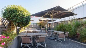 An outdoor patio with multiple tables and chairs, surrounded by greenery and a large umbrella providing shade. - B&B Hotel Toulouse Université (Toulouse)