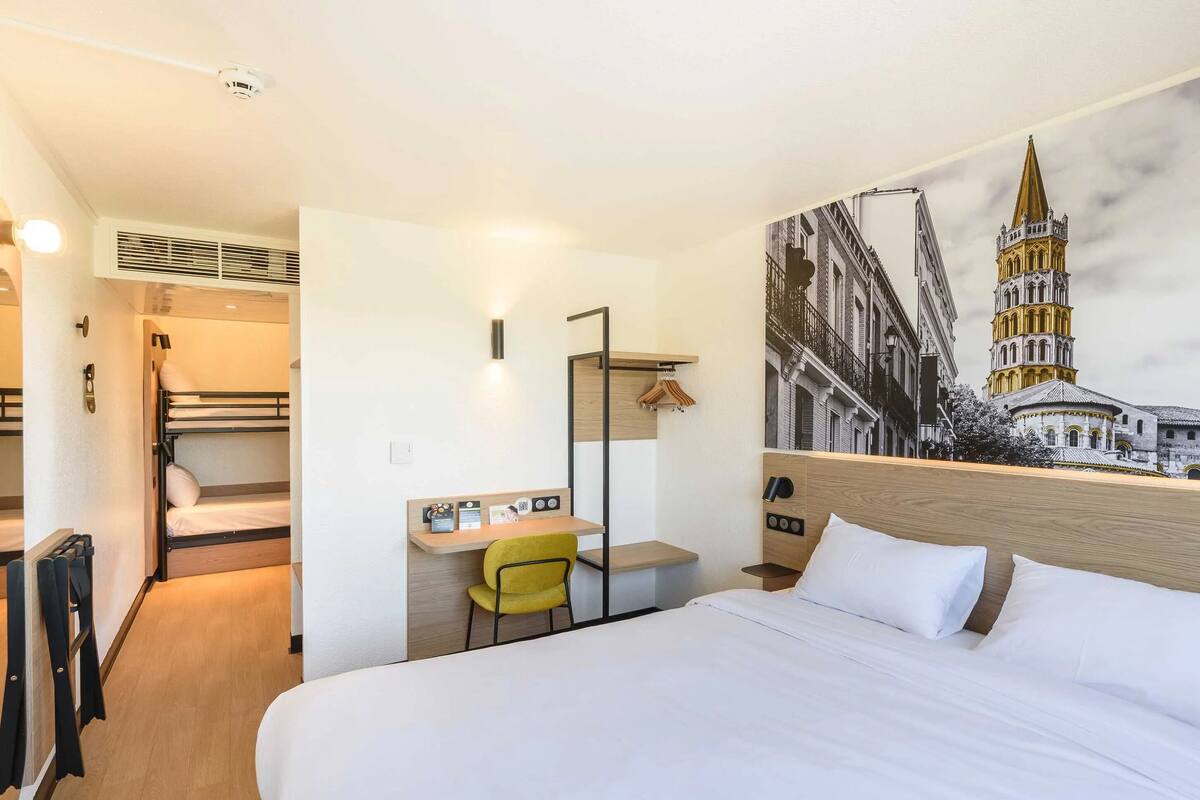 A hotel room with a bed, a desk with a chair, a coat rack, and a wall-mounted lamp.