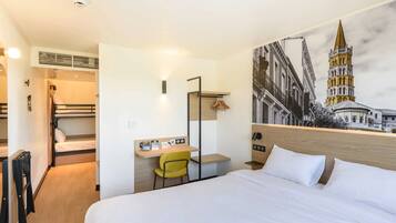 Habitación de hotel con cama, escritorio, silla, perchero y lámpara de pared.