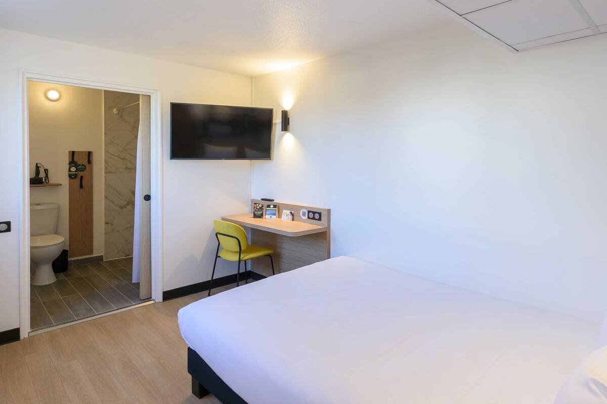 A hotel room with a bed, a desk with a chair, a television mounted on the wall, and a bathroom visible through an open door.