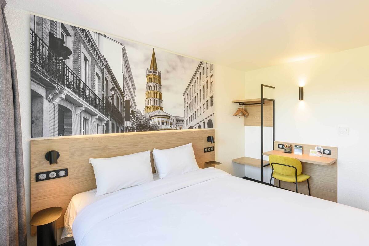 A hotel room with a bed, a desk, a chair, a lamp, and a wall-mounted TV.