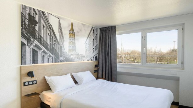 A hotel room with a bed, a nightstand, a wall-mounted light, a window with curtains, and a view of buildings. - B&B Hotel Toulouse Université (Toulouse)