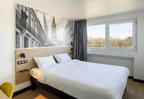 A hotel room with a bed, a nightstand, a wall-mounted light, a window with curtains, and a view of buildings. - B&B Hotel Toulouse Université (Toulouse)