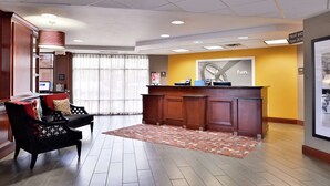 A hotel lobby with a reception desk, a seating area with a patterned rug, and a large window with curtains.