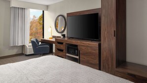 A hotel room with a large bed, a flat-screen TV, a wooden entertainment unit, a desk with a chair, and a window with curtains.