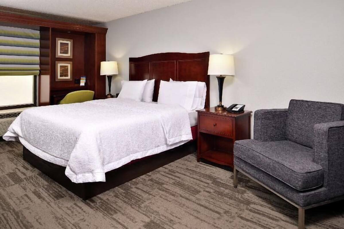 A hotel room with a large bed, two bedside lamps, a gray armchair, and a wooden nightstand.