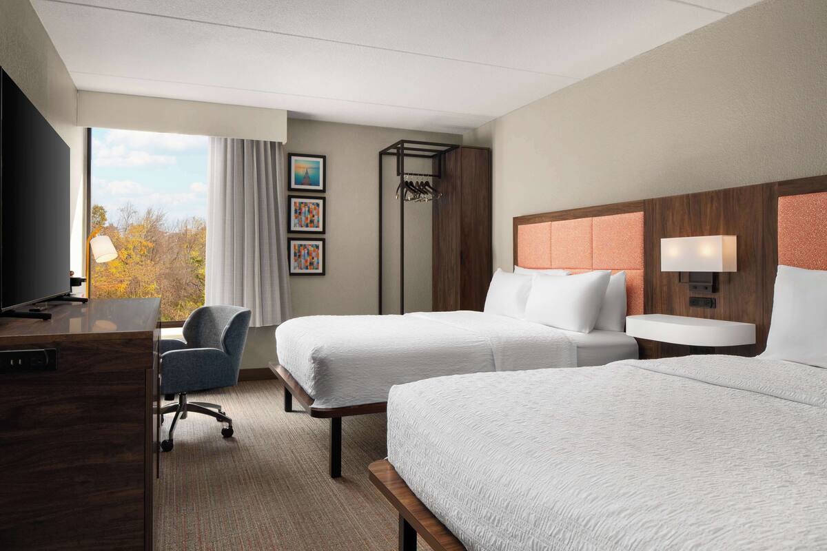 A hotel room with two beds, a desk, a chair, a TV, and a window with a view of trees.