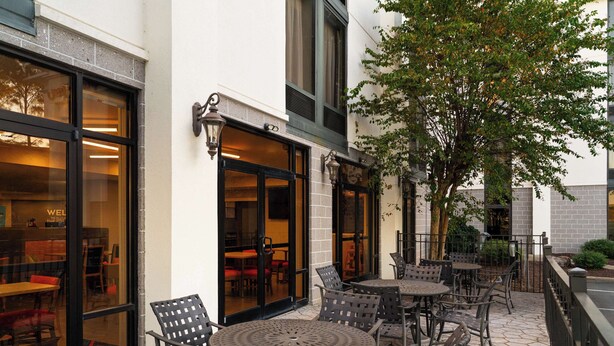 An outdoor patio with tables and chairs, a tree, and a building with large windows and doors.