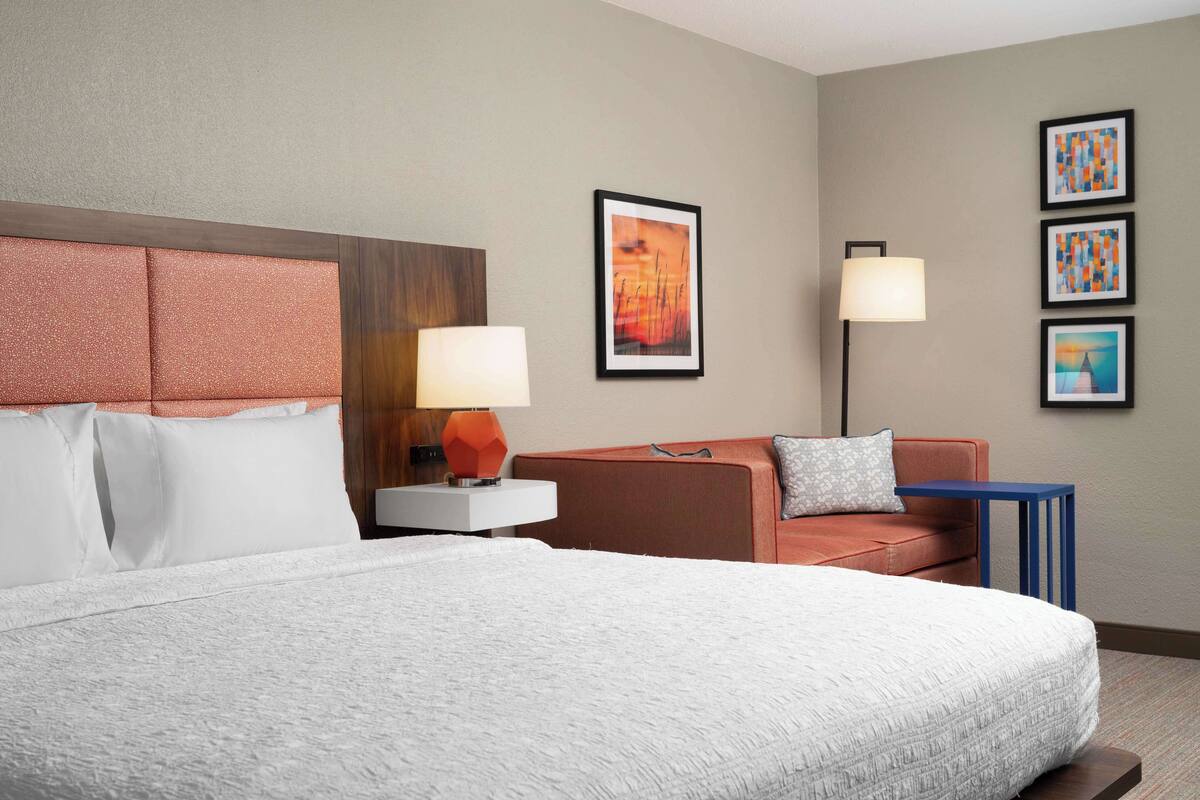 A hotel room with a large bed, a sofa, a nightstand with a lamp, and framed artwork on the wall.