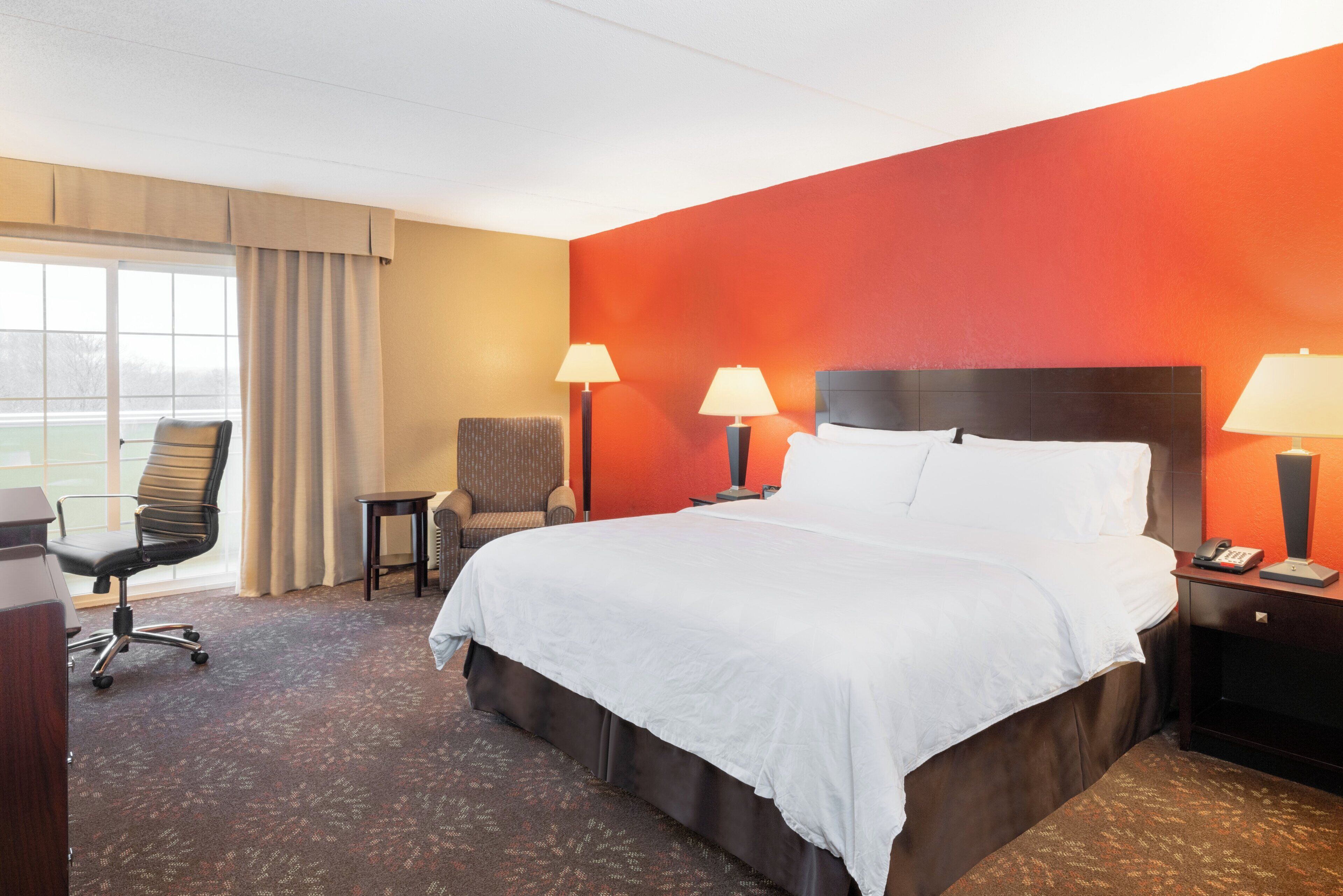 A hotel room with a large bed, two bedside lamps, a chair, a desk, and a view through a window.