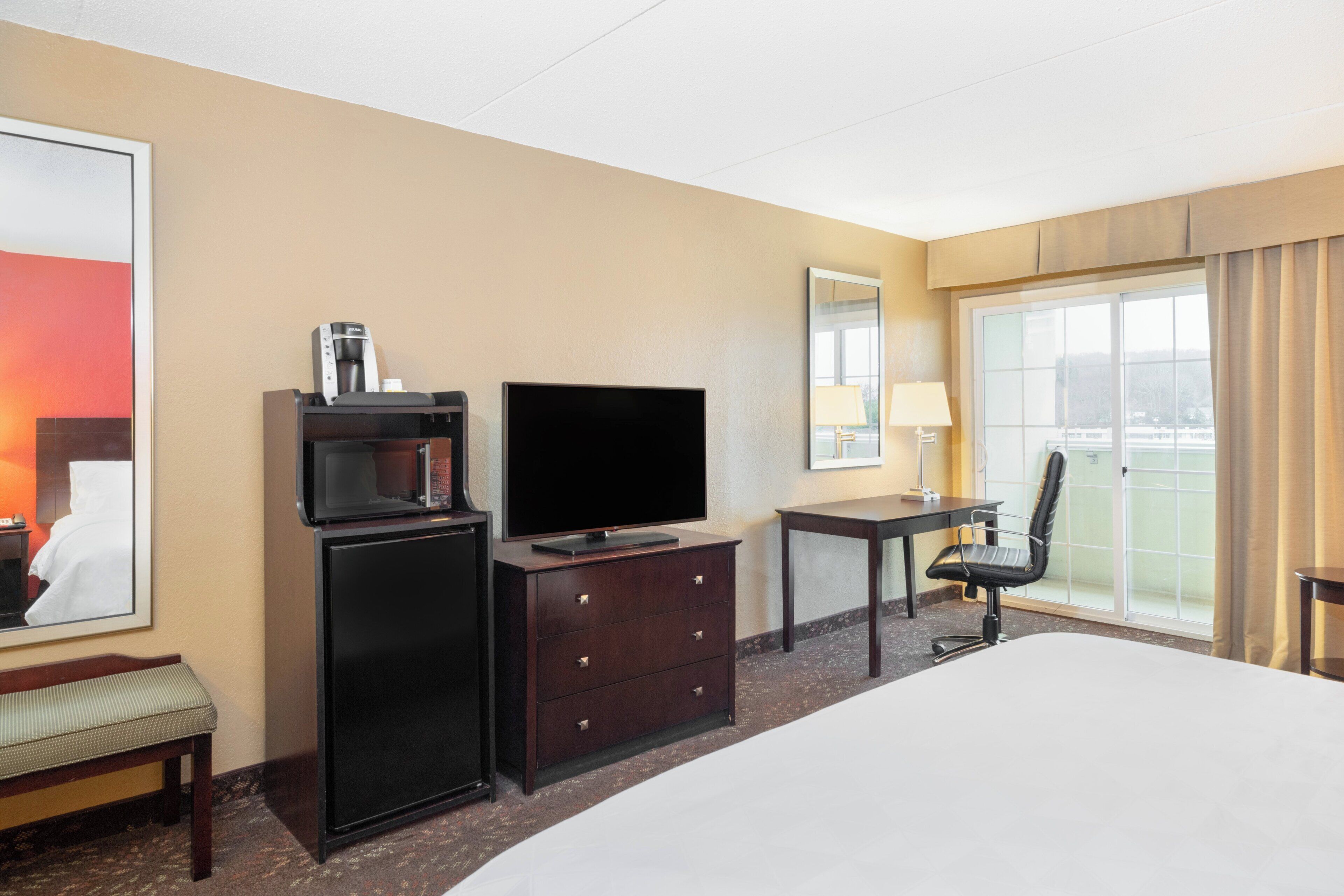 Habitación de hotel con cama, escritorio con silla, televisor, minibar y una ventana grande con cortinas.
