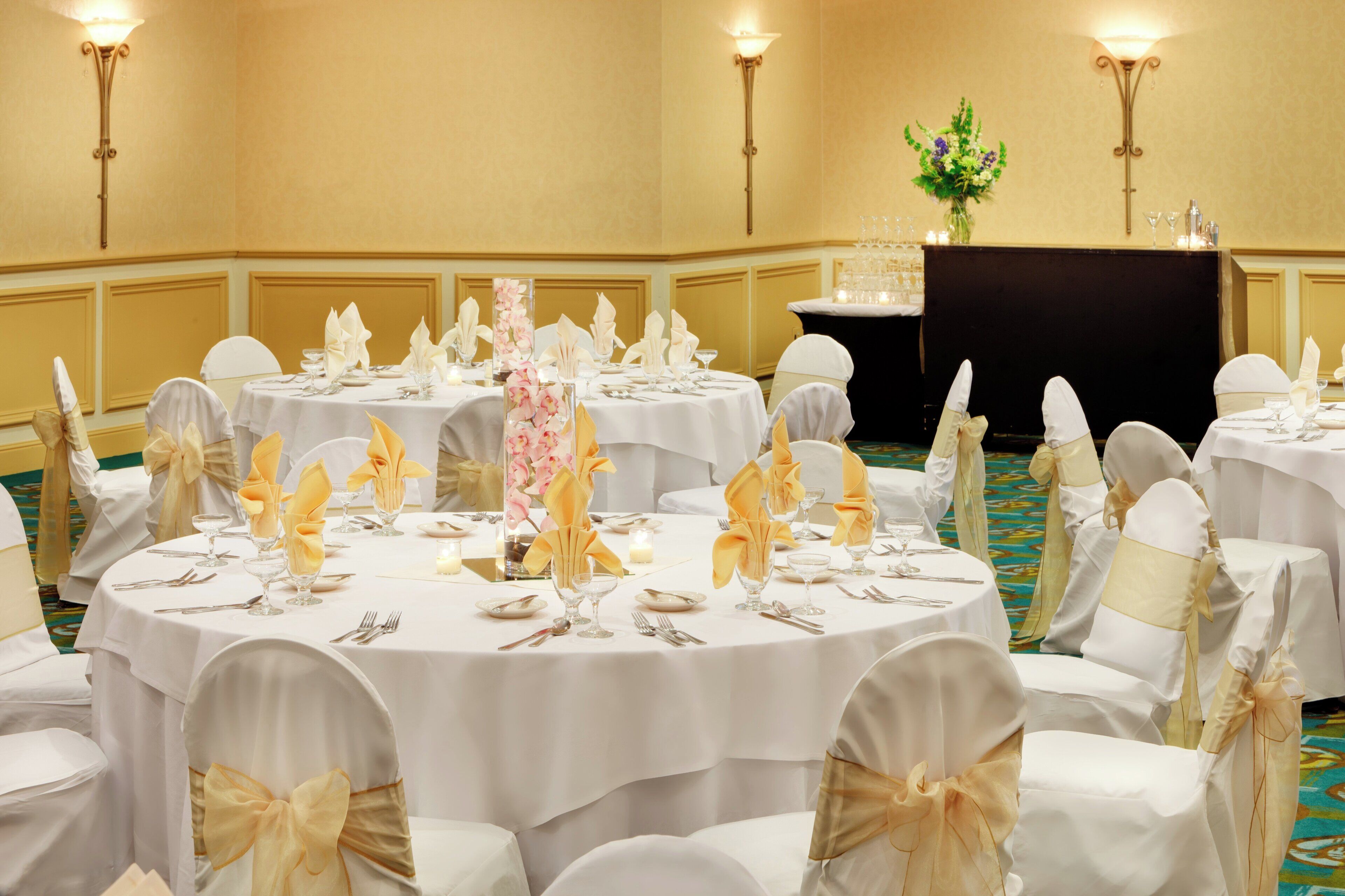 Een balzaal met ronde tafels gedekt voor een formeel evenement, met witte tafelkleden, gouden linten en bloemstukken als middelpunt.