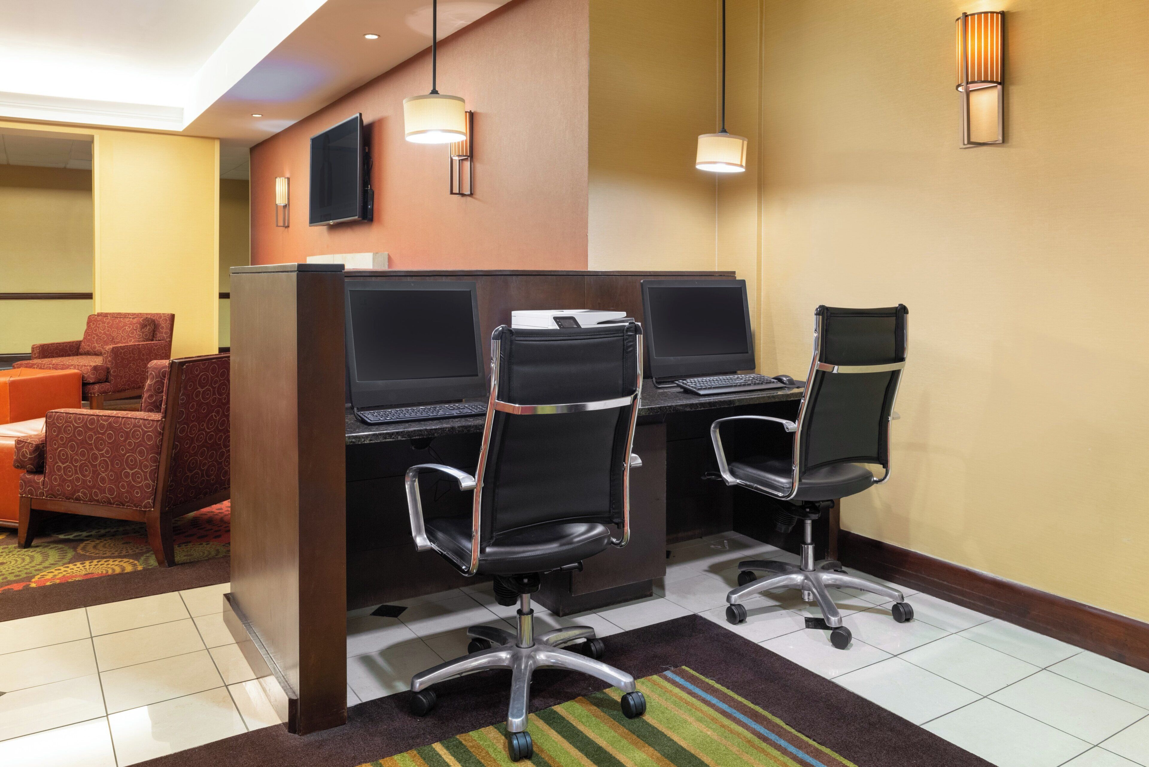 A hotel room with a desk area featuring two computer workstations, a television mounted on the wall, and a seating area with two armchairs.