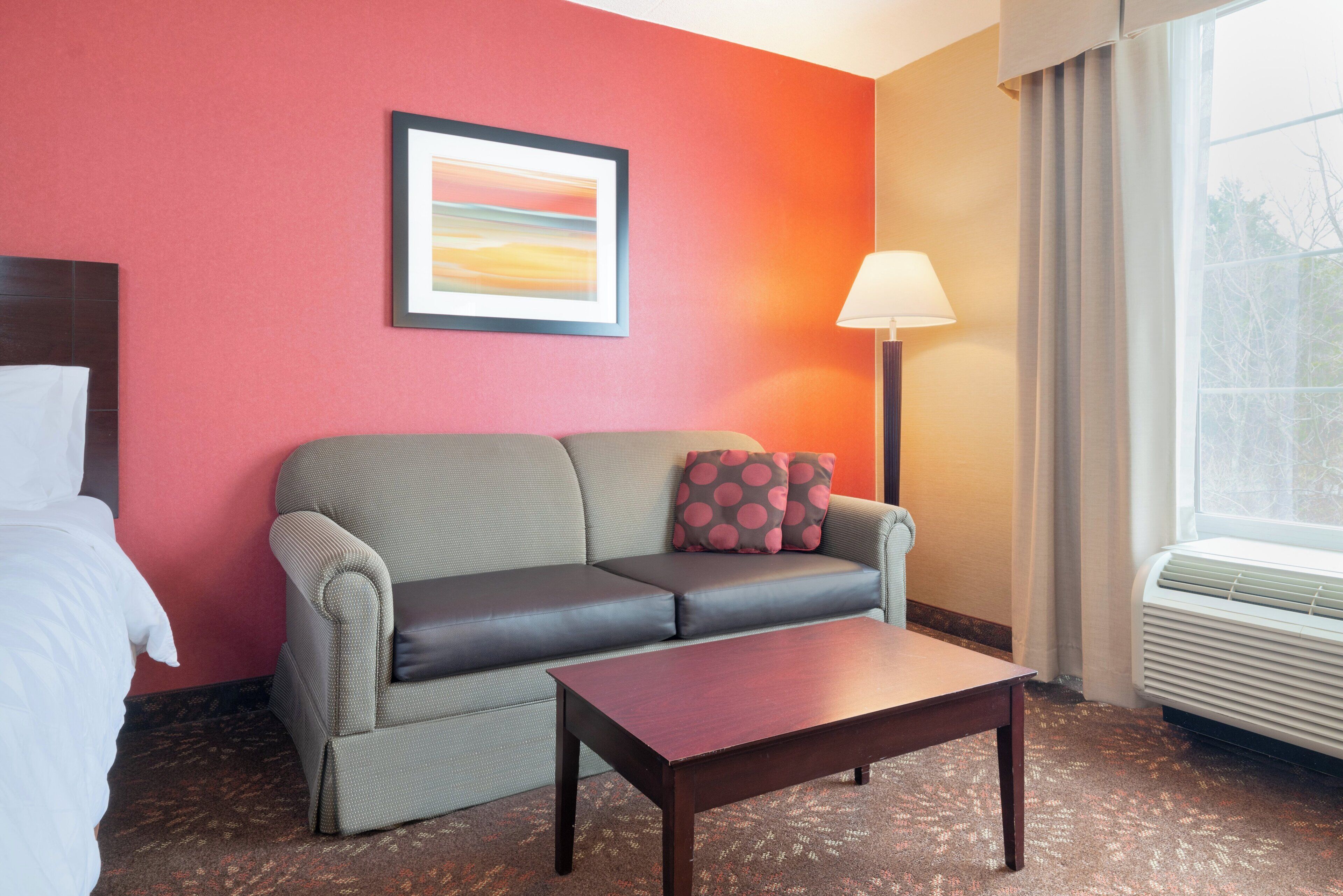 A hotel room with a sofa, a small table, a lamp, and a window with curtains.