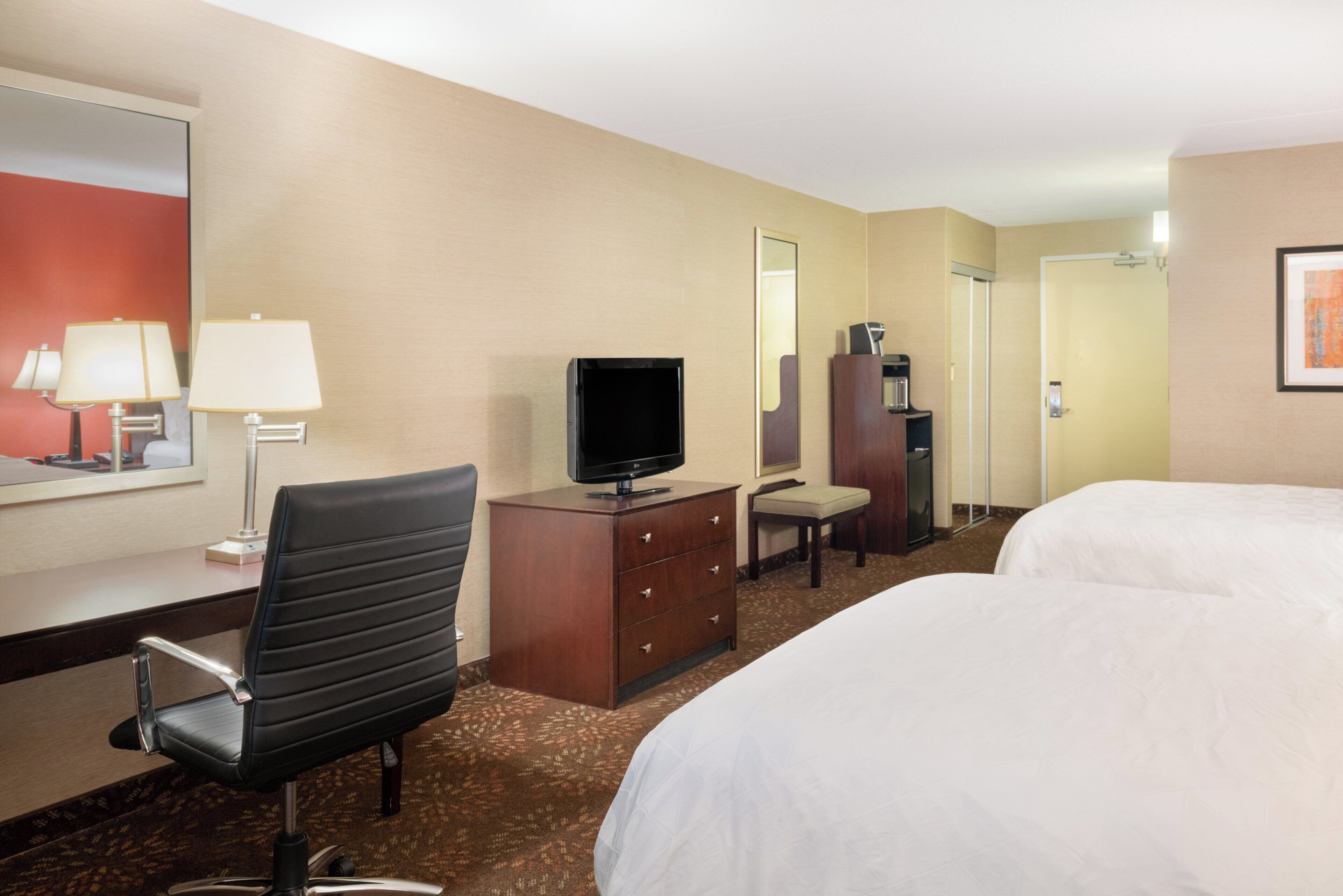 A hotel room with a bed, a desk with a lamp, a television, a chair, a dresser, and a mirror.