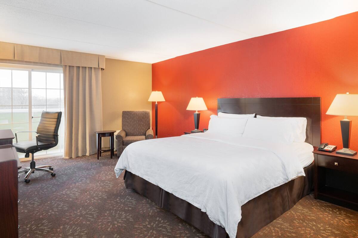 A hotel room with a large bed, two bedside lamps, a chair, a small table, and a view of a tennis court through the window.